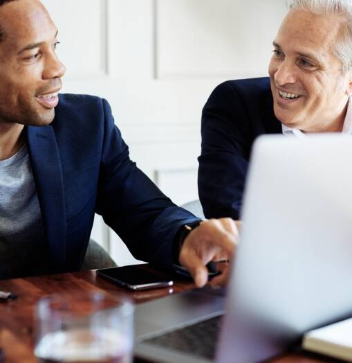 Two men in business attire sit at a table, smiling and having a discussion while looking at a laptop screen. One man points at the laptop, suggesting a collaborative or friendly work environment.