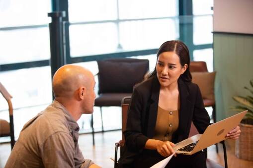 A woman holding a laptop talks to a man in an office setting. Both are seated and appear to be engaged in discussion, with chairs and large windows in the background.