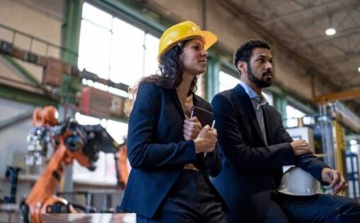 Two professionals in business attire, one wearing a yellow hard hat, sit together in a large industrial facility with robotic arms in the background, appearing thoughtful and attentive.