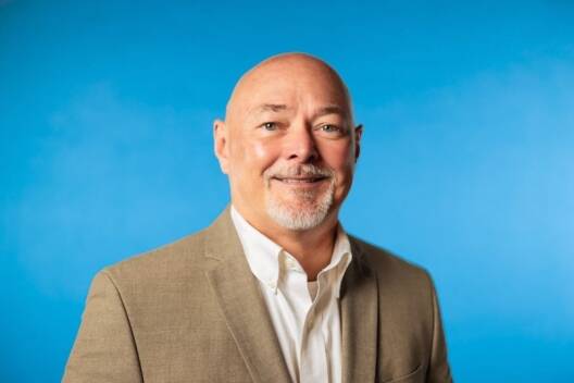 A middle-aged man with a bald head and gray goatee smiles while wearing a beige blazer and white shirt, standing against a plain blue background—an ideal image for Lean Manufacturing Consulting professionals.
