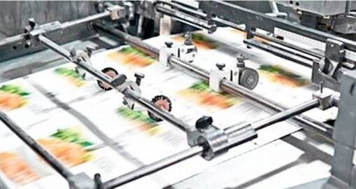 A close-up of a printing press in operation, producing colorful printed sheets with images of vegetables. Metal rollers and guides ensure precise movement—an example of Continuous Improvement in manufacturing processes.