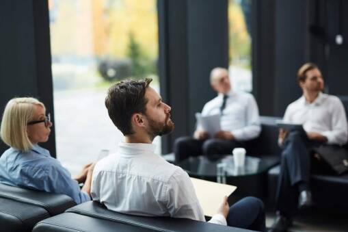 Four people in business attire sit on black couches in a modern, glass-walled office, attentively listening and holding papers. The background shows trees and natural light coming through large windows.