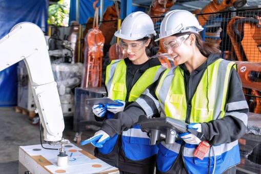 Two women wearing safety helmets, vests, and gloves operate a robotic arm in an industrial setting. They hold control devices and smile while monitoring the robotic equipment. Machinery and safety fencing are visible in the background.